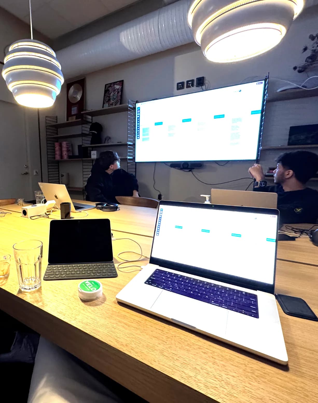 Team members working at a shared table with laptops and a wall-mounted project board.